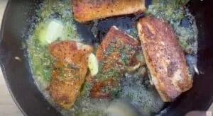 Pan-seared salmon fillets sizzling in lemon herb butter sauce, cooked in a black cast-iron skillet.